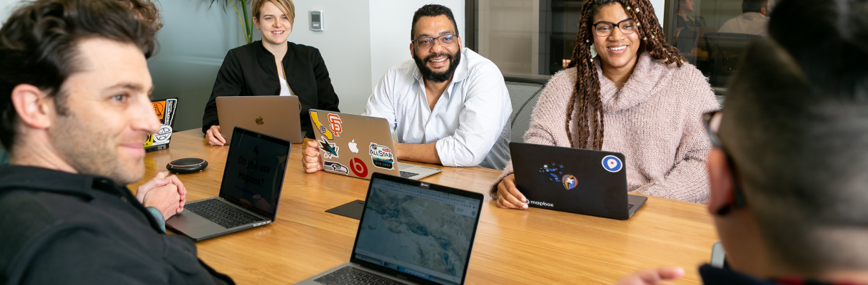 6 Pessoas diversas reunidas ao redor de uma mesa conversando com sorriso no rosto